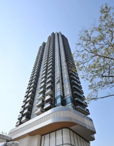 Tall glass skyscraper with stacked balconies stretching upward against a clear blue sky a tree visible on the right