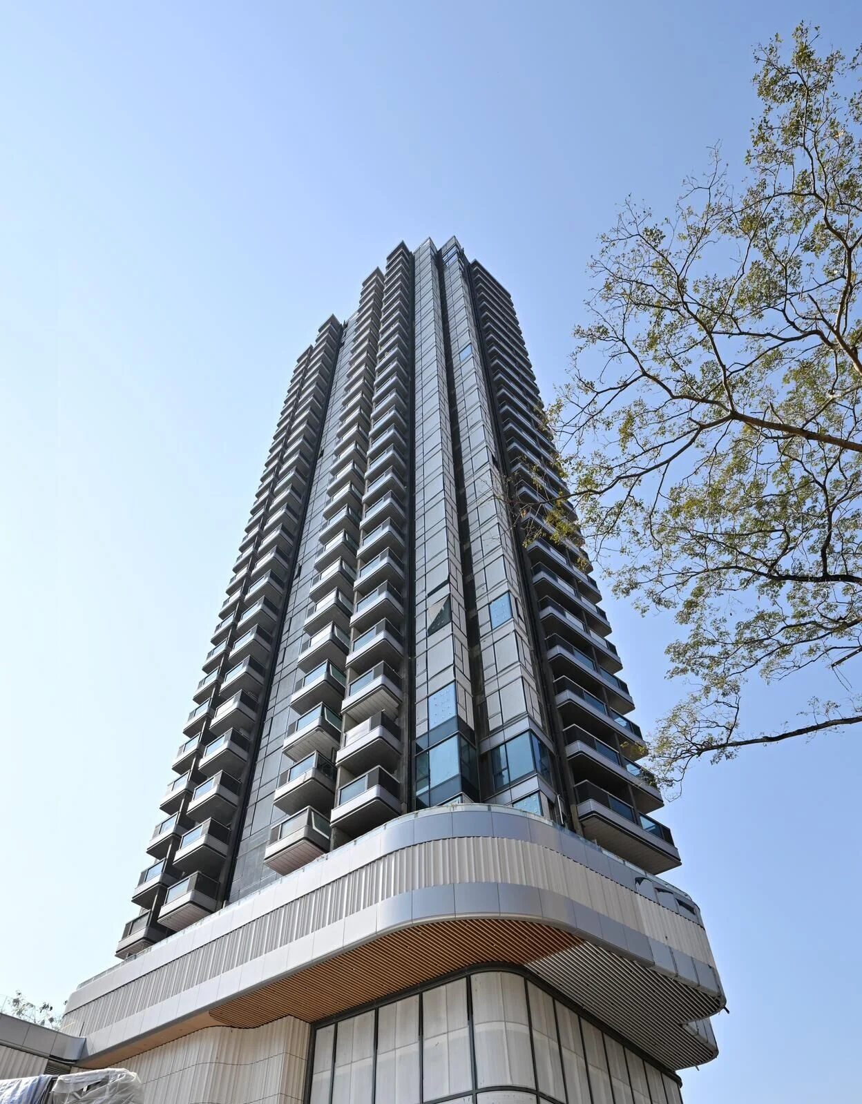 Tall glass skyscraper with stacked balconies stretching upward against a clear blue sky, a tree visible on the right.