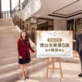 Two staff members in business attire pose beside a large signboard in a marble hotel lobby with a curved staircase behind them