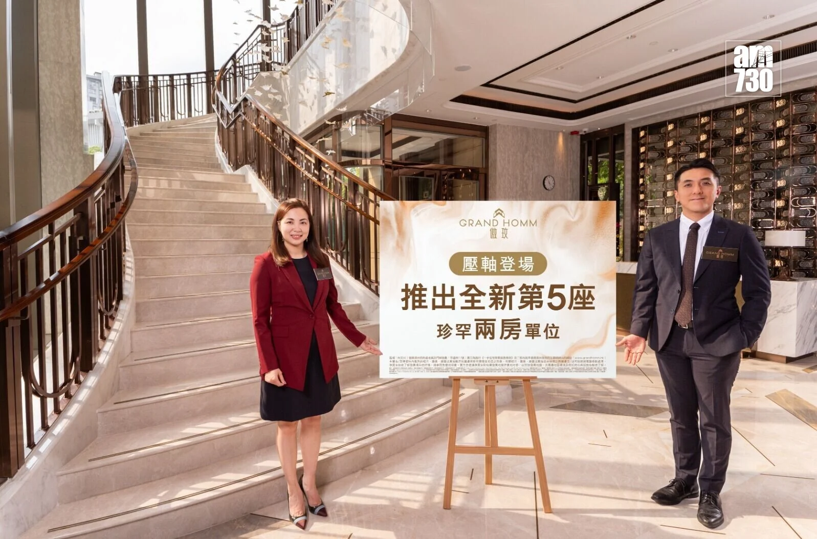 Two staff members in business attire pose beside a large signboard in a marble hotel lobby with a curved staircase behind them.