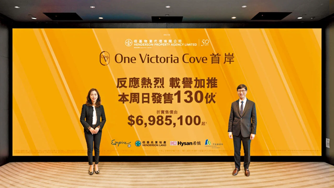 Two formally dressed presenters stand on either side of a large orange One Victoria Cove banner with Chinese text and the price $6,985,100.