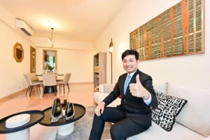 Man in a suit giving a thumbs up while seated on a beige sofa in a modern living room with a dining area in the background