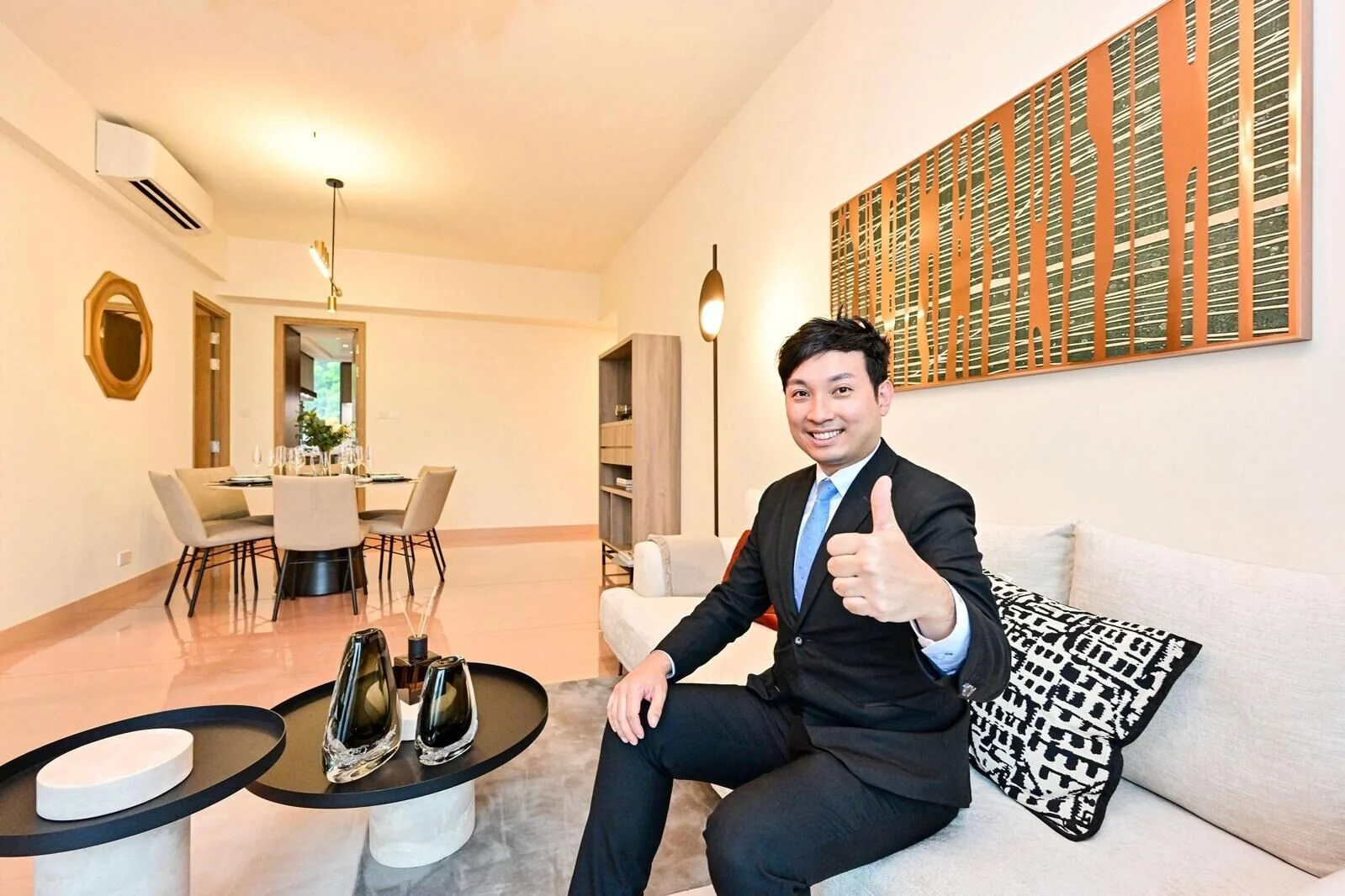 Man in a suit giving a thumbs up while seated on a beige sofa in a modern living room with a dining area in the background.