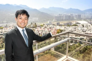 Man in a dark suit and teal tie stands on a balcony thumbs up with a cityscape and mountains in the background