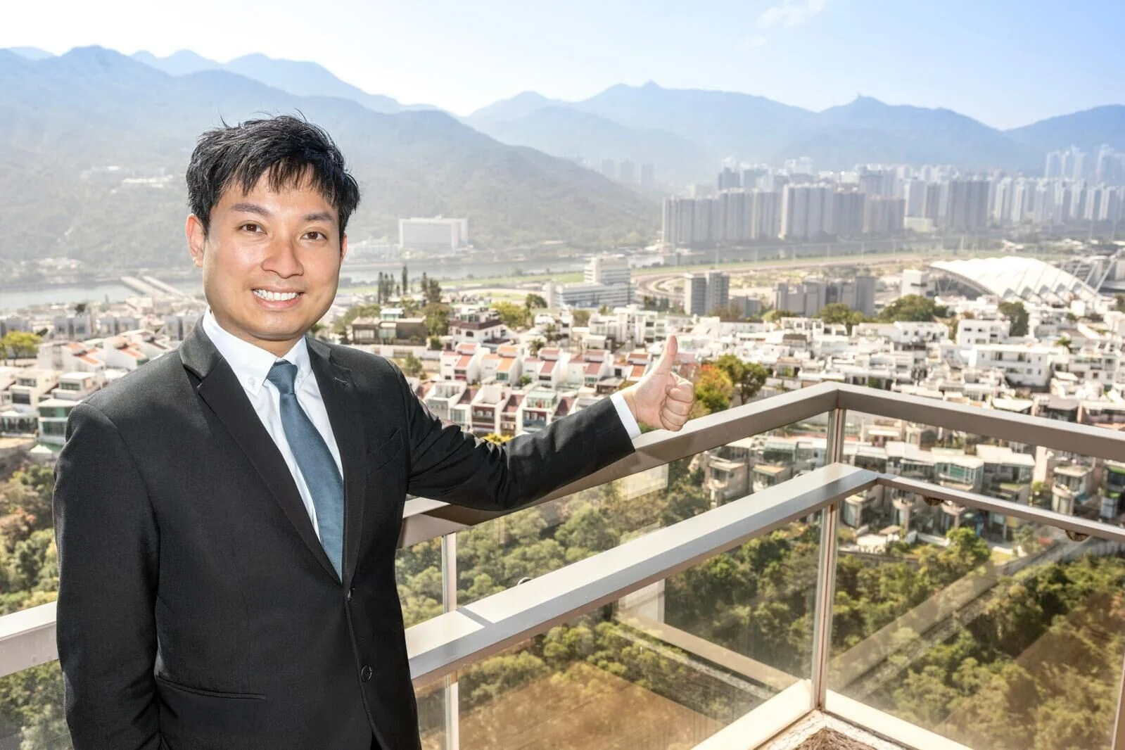 Man in a dark suit and teal tie stands on a balcony, thumbs up, with a cityscape and mountains in the background.
