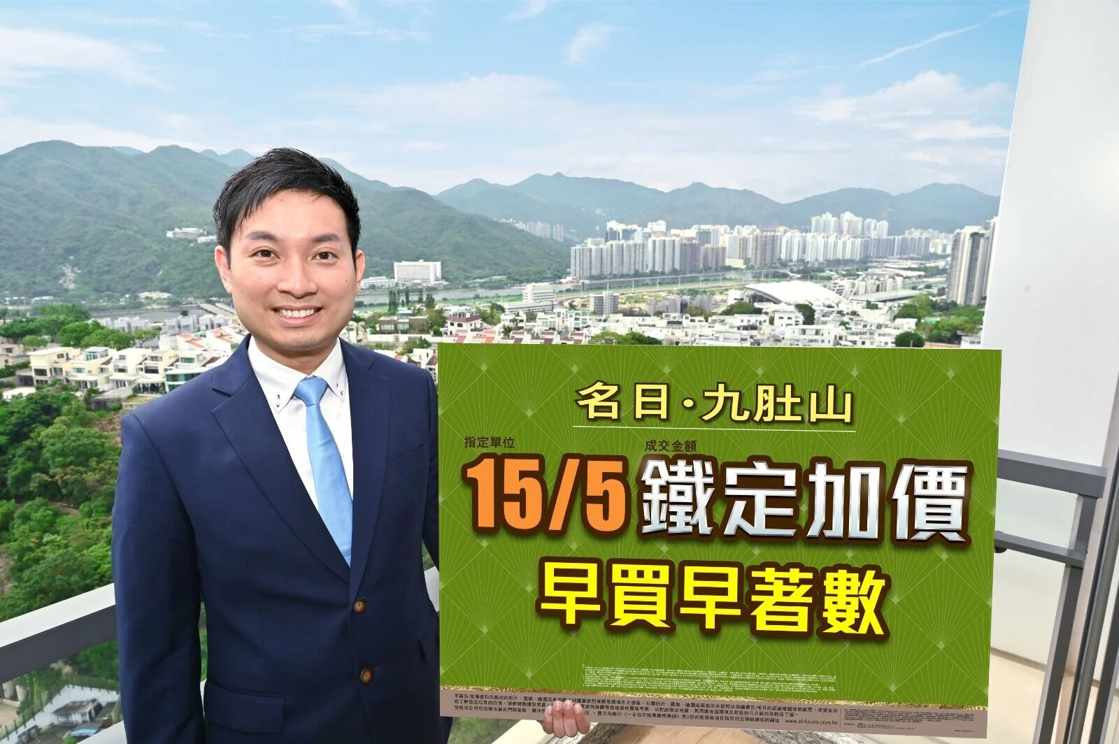 Asian man in a navy suit on a balcony, holding a large green sign with Chinese text; city skyline and mountains in the background.