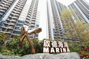 Bronze abstract sculpture in foreground with tall glass residential towers and a large sign reading MARINA in Chinese and English behind landscaped plants