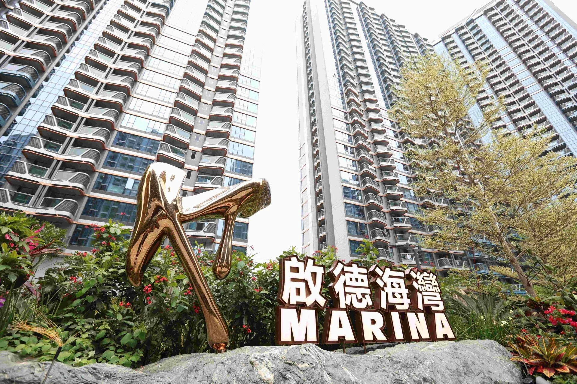 Bronze, abstract sculpture in foreground with tall glass residential towers and a large sign reading MARINA in Chinese and English behind landscaped plants.