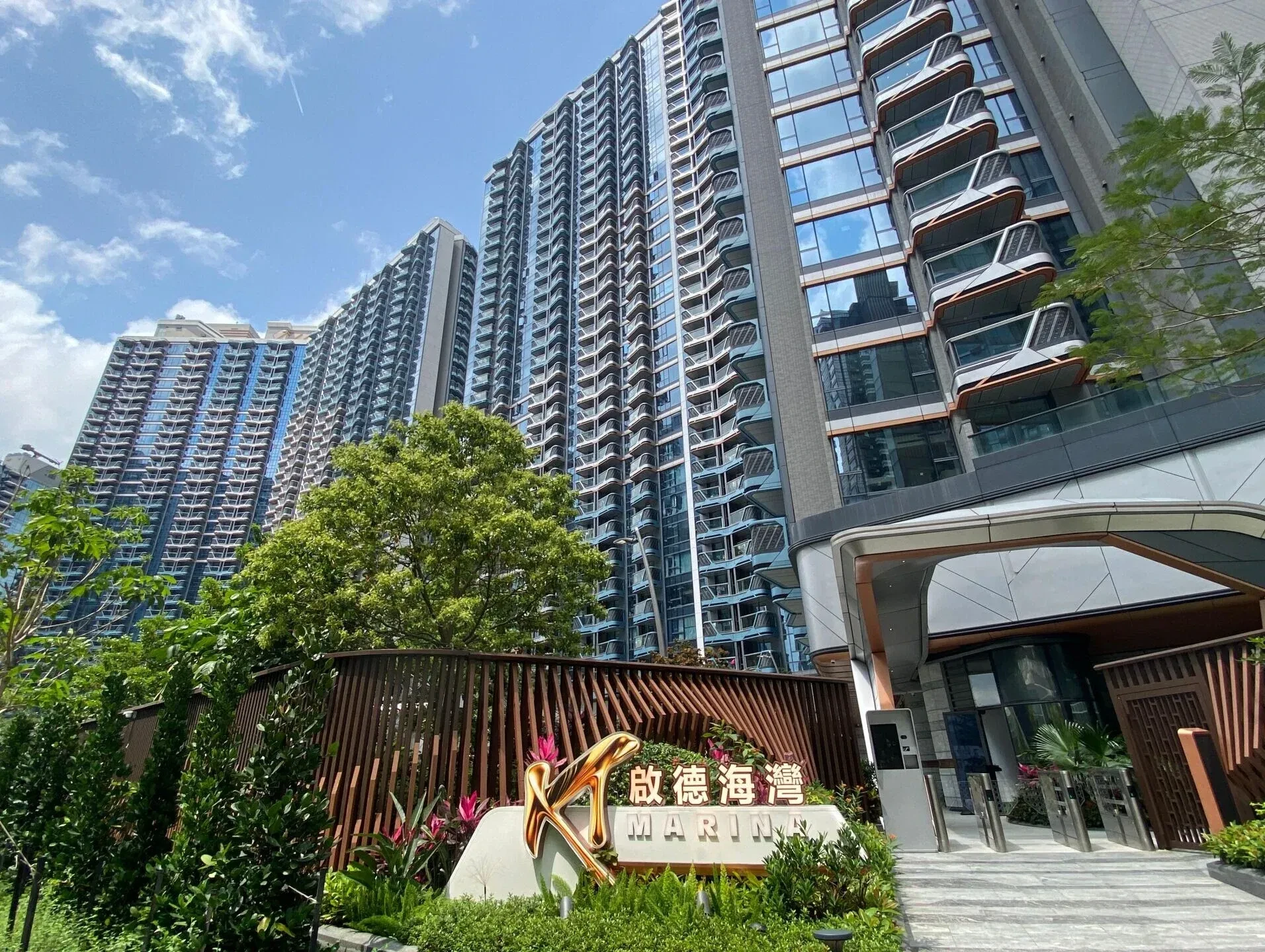Tall modern high-rise buildings with glass balconies against a blue sky, foreground includes a sign reading 'MARINA' with greenery.
