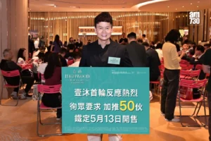 Man in a black suit stands at a busy event holding a teal promotional board for Highwood Phase 2 in the foreground In the background attendees sit and mingle a logo is visible in the top right corner
