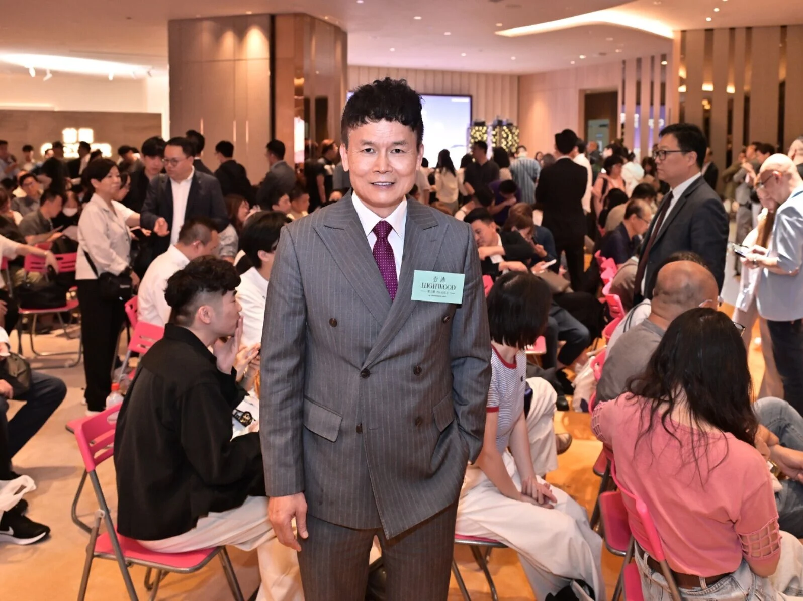 Man in a gray pinstripe suit with a name badge stands at the center of a crowded event hall filled with seated attendees and crowds in the background.