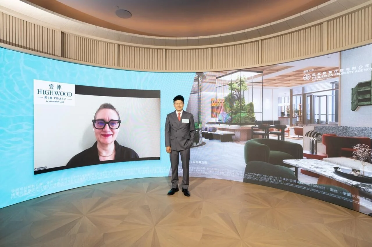 Man in a gray suit stands in a curved showroom backdrop; a video call on the left shows a woman with glasses, while a living-room scene fills the right.