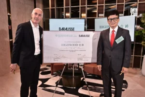 Two men in suits pose with a large certificate labeled HIGHWOOD in a modern lobby setting