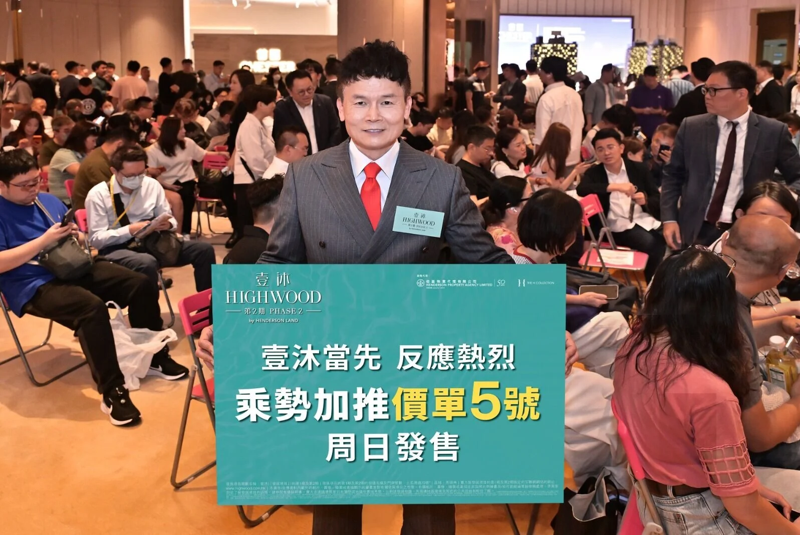 Man in a gray pinstripe suit and red tie stands at a busy event, holding a teal banner about Highwood Phase 2 as attendees chat in the background.