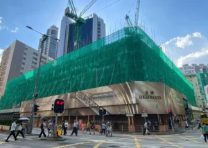 Under construction building wrapped in green safety netting with cranes overhead pedestrians cross a busy street below