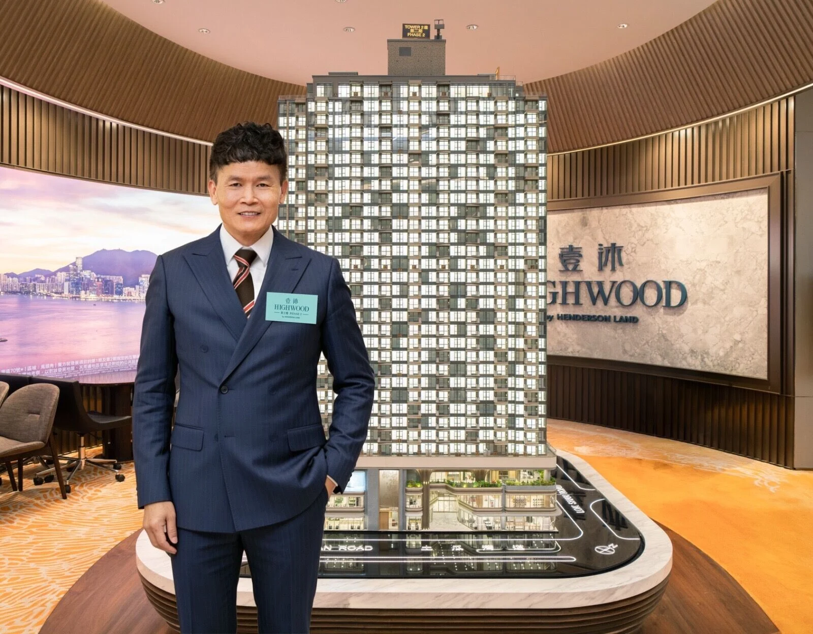 Smiling man in a navy suit stands beside a tall apartment building model in a showroom, with a 'HIGHWOOD' sign on the wall behind him.