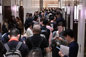 Crowded elevator lobby with dozens of people standing in a narrow hallway between elevator doors