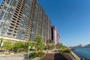 Row of glass fronted high rise apartment buildings along a waterfront promenade with trees and a paved path under a clear blue sky