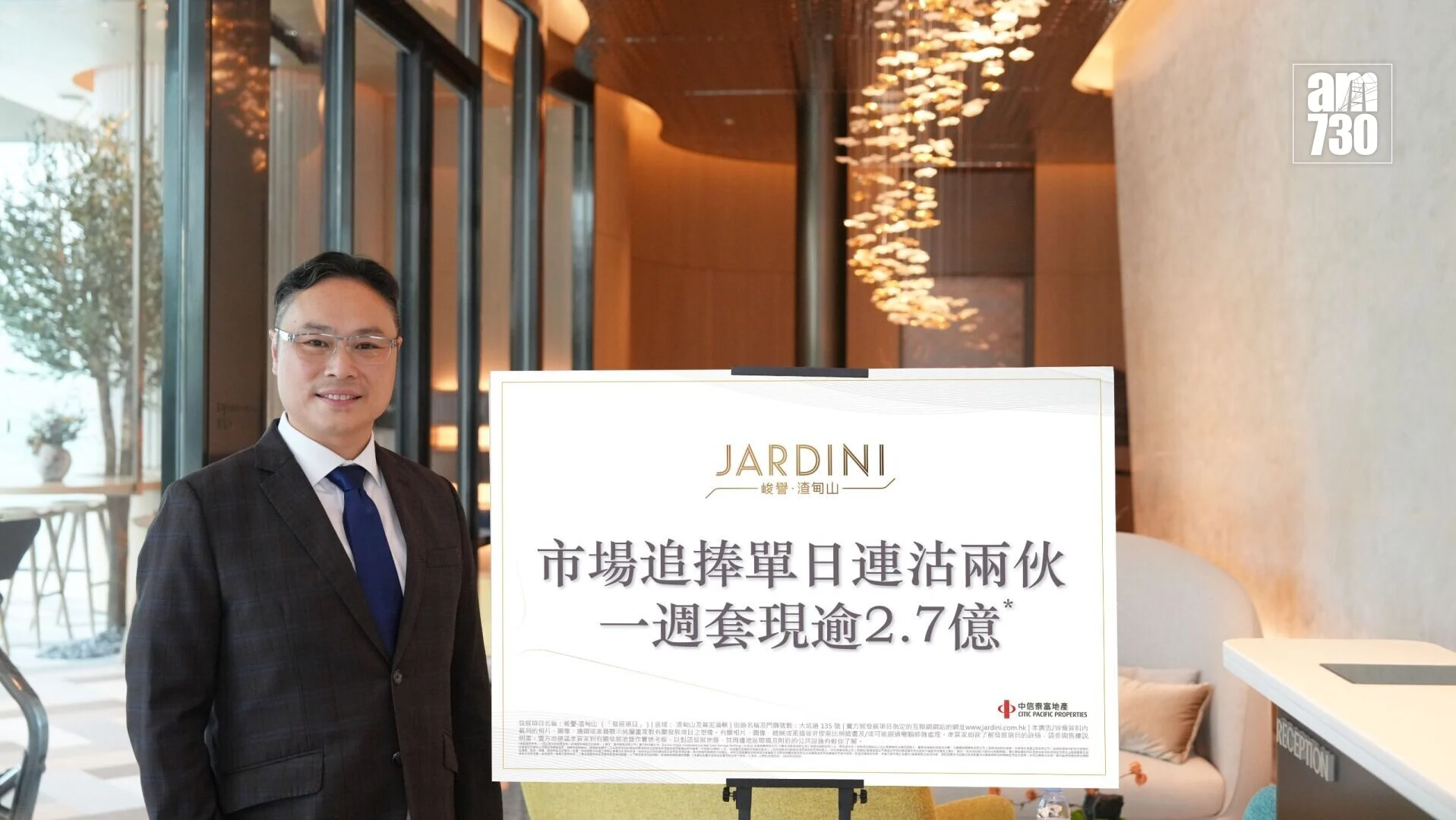Asian man in a dark suit and blue tie smiling beside a large white poster on an easel in a modern, well-lit lobby with decorative lights in the background.