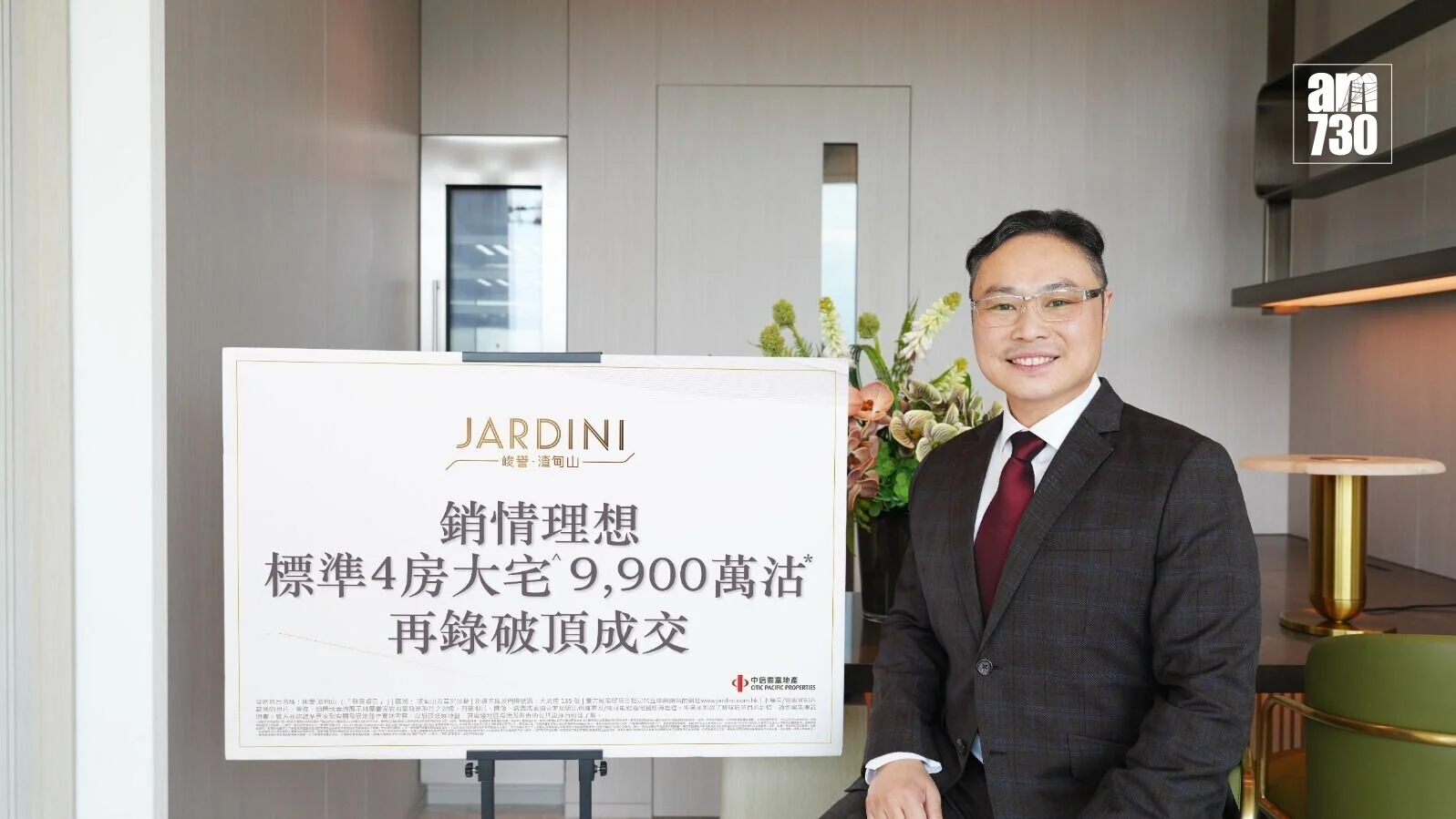 Man in a dark suit and red tie smiling beside a large JARDINI promotional sign in a modern lobby setting