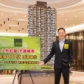 Asian man in a dark suit with a yellow Lime badge stands beside a tall high rise model and a green advertisement board in a showroom