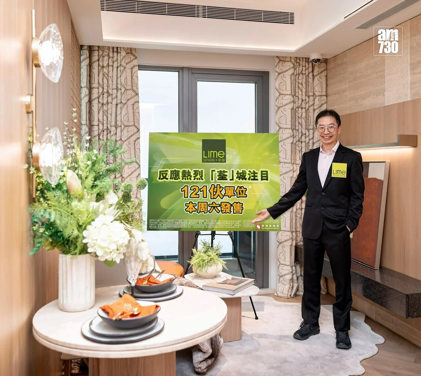 Man in a dark suit posing beside a lime-green promotional sign in a showroom setting, smiling at the camera with a floral arrangement nearby (LIME Spark text on sign).