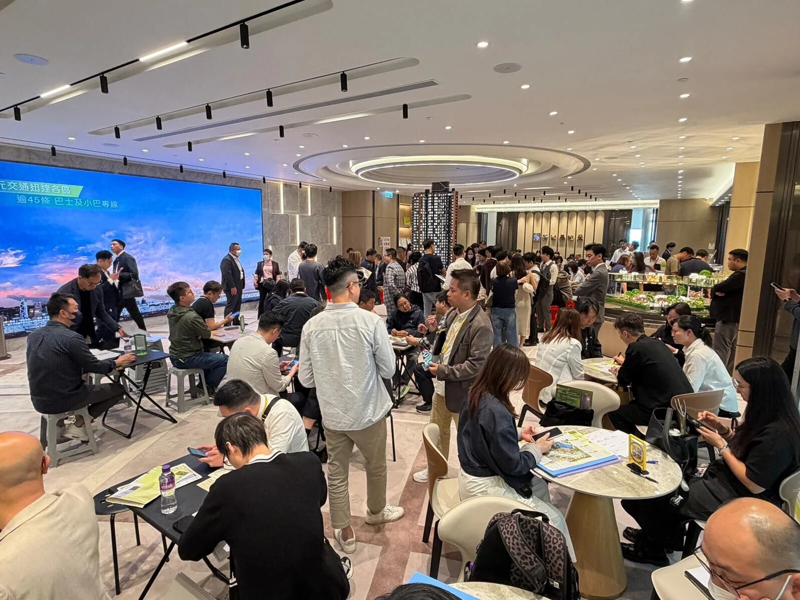Crowded business networking event in a modern hall: people sit at round tables, chat, and review materials near a large blue presentation screen and buffet area.