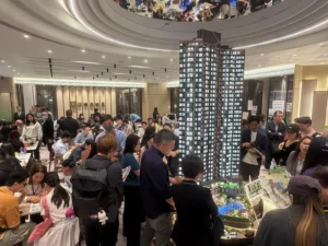 Crowded real estate sales showroom around a large scale model of a tall apartment building with people examining brochures and talking with agents