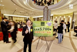 Smiling man in a suit gives a thumbs up beside a large green sign announcing 5700 units sold at an indoor real estate event Crowds are gathered in the background