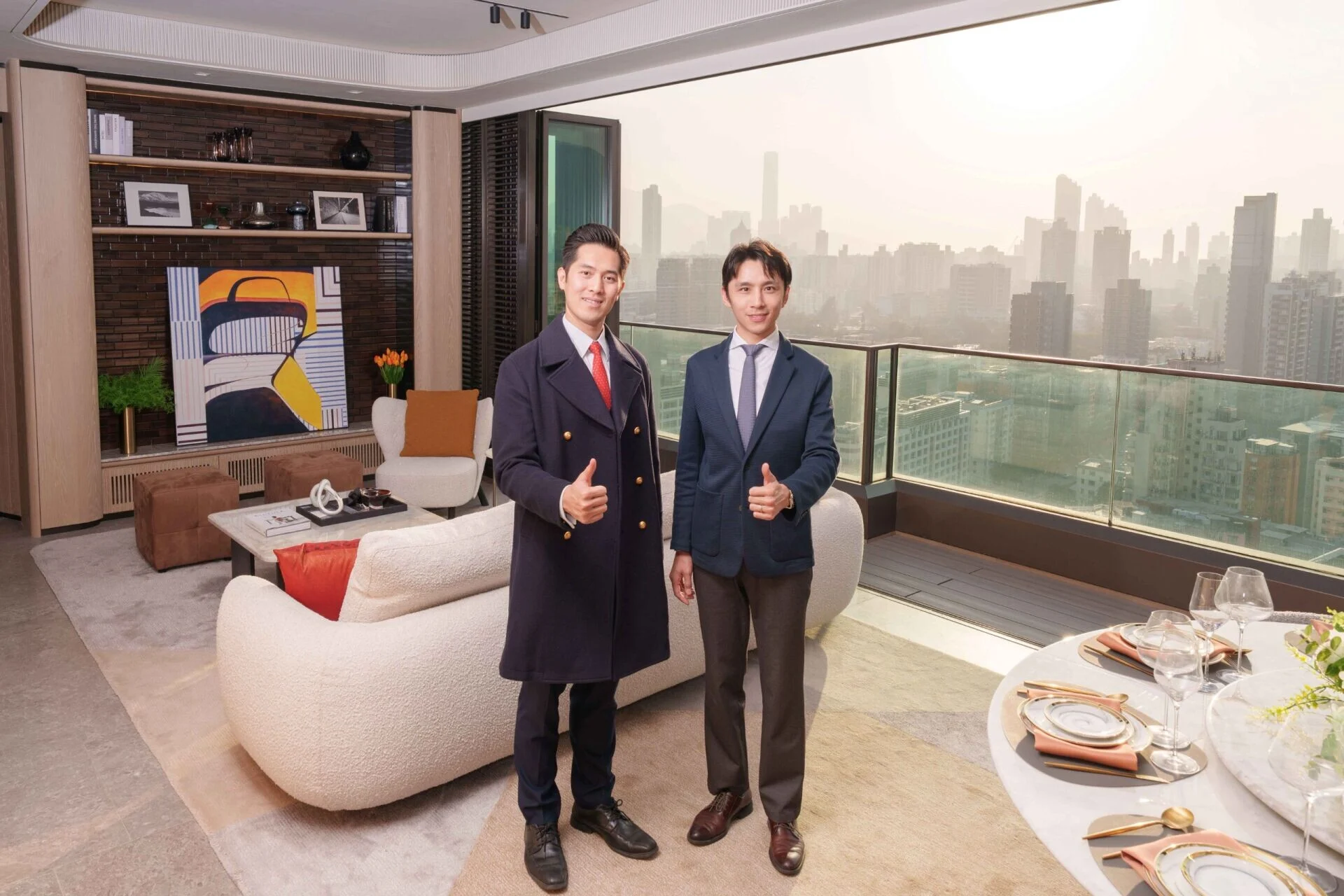 Two professionally dressed men stand in a modern living room with a city skyline view, giving thumbs up to the camera.