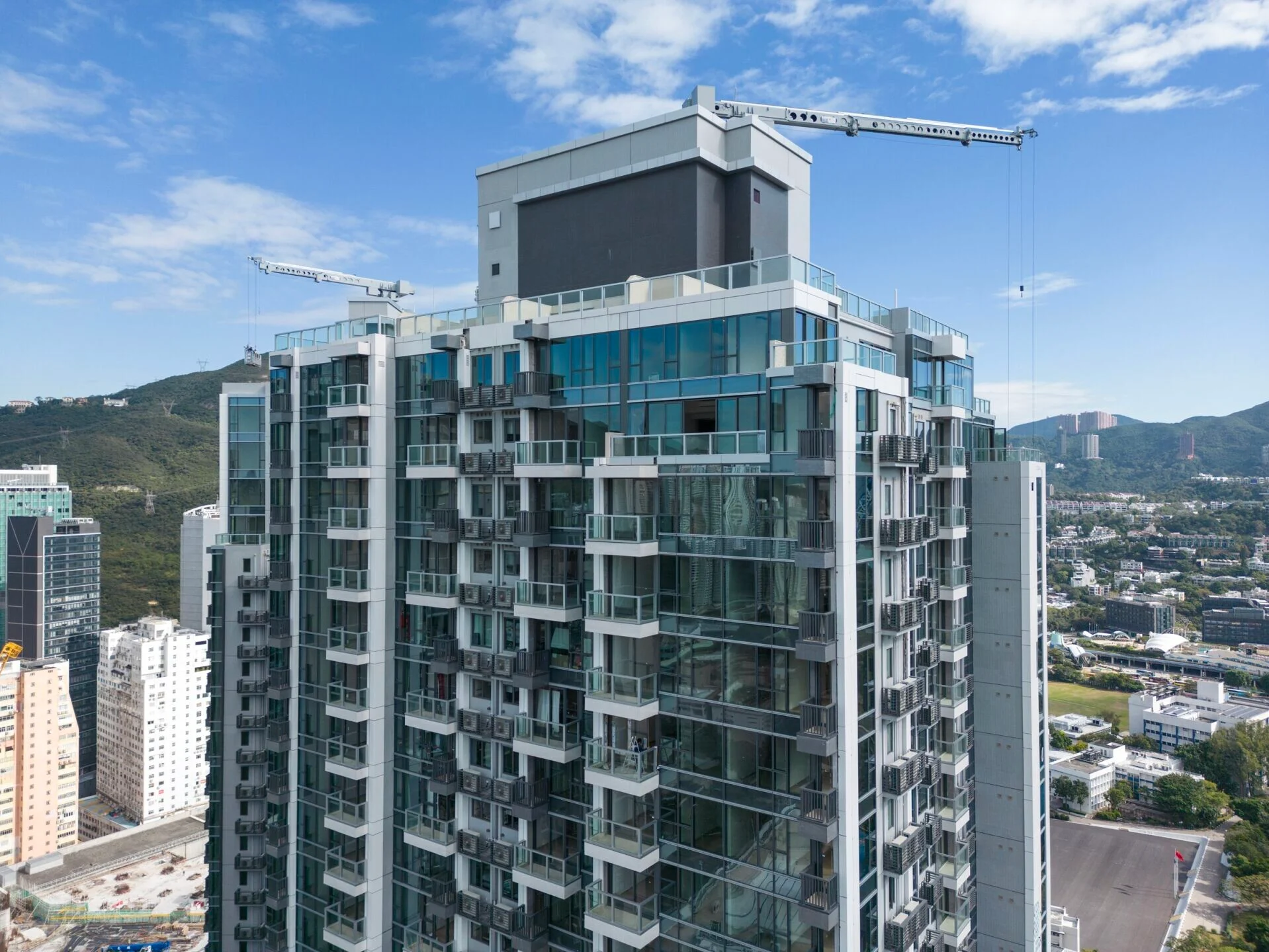 Modern glass apartment building under construction with cranes against a blue sky, skyline in the background.