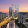 Twin glass skyscrapers dominate a city skyline at sunset beside a river with a curved highway and surrounding high rise apartments AM730 logo in the top right corner