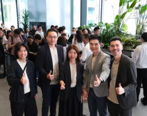Five coworkers in business casual pose and thumbs up in a bright office lobby with plants and a crowded background