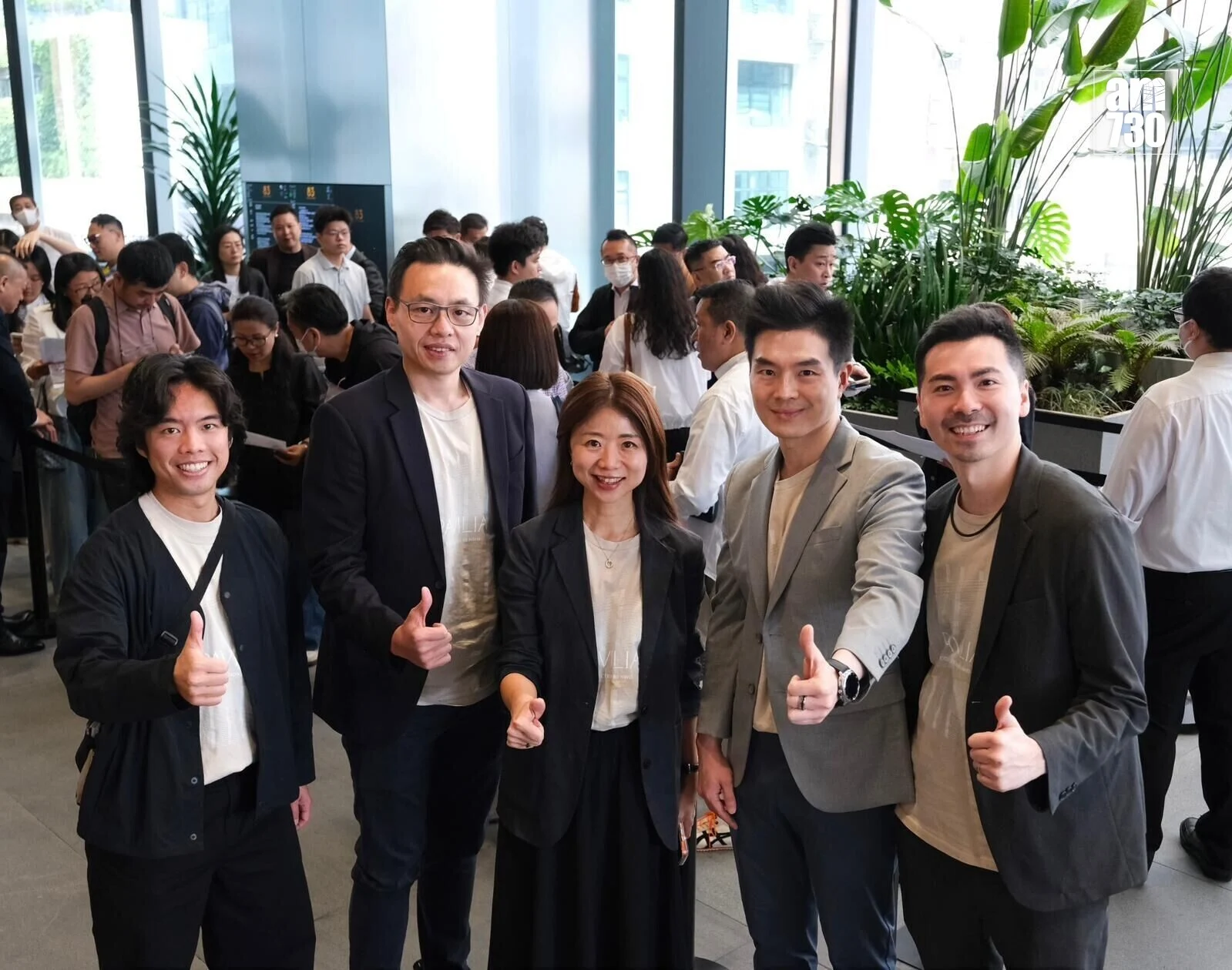 Five coworkers in business casual pose and thumbs up in a bright office lobby, with plants and a crowded background.