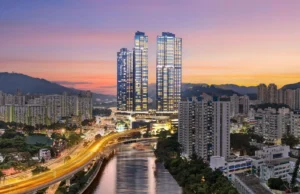 Twin illuminated skyscrapers rise over a city at dusk with a sunset sky and a winding canal below