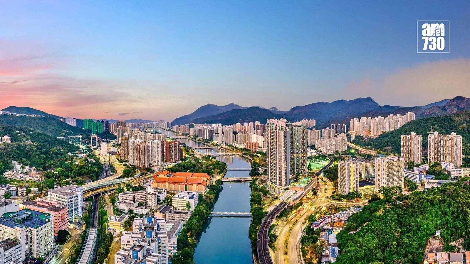 Panoramic cityscape with a river winding through high-rise buildings and green hills, under a pastel sunset; am730 logo in the upper right corner.