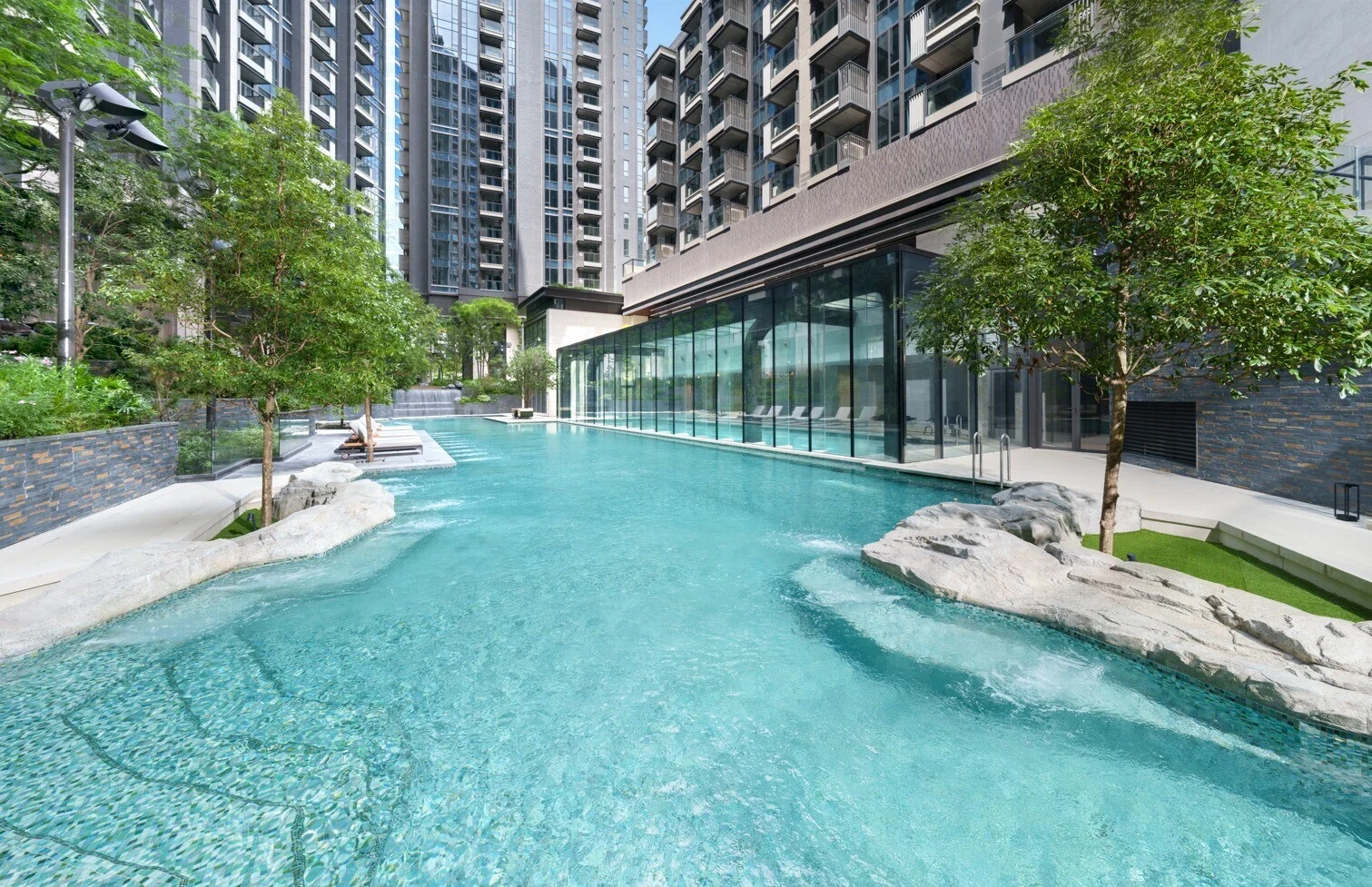Outdoor pool area at a high-rise residential complex, with glass-walled building and turquoise water surrounded by trees and stone features.