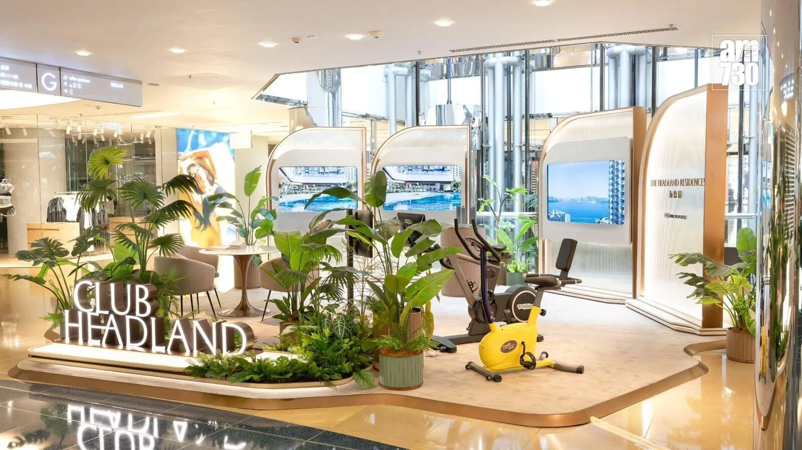 Indoor showroom with lush plants, a yellow stationary bike, and a illuminated 'Club Headland' sign near display screens.