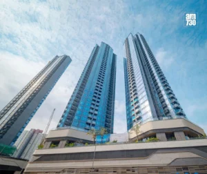 Low angle view of three tall glass skyscrapers reaching into a blue sky with a curved beige podium at street level and an AM730 logo in the top right corner