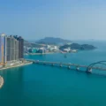 Aerial view of a coastal city tall colorful apartment towers beside a long bridge over blue green water with hills in the distance and a logo in the top right corner
