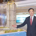 Man in a dark suit and red tie stands beside a large architectural scale model of tall waterfront towers in a showroom