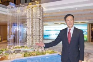 Man in a dark suit and red tie stands beside a large architectural scale model of tall waterfront towers in a showroom