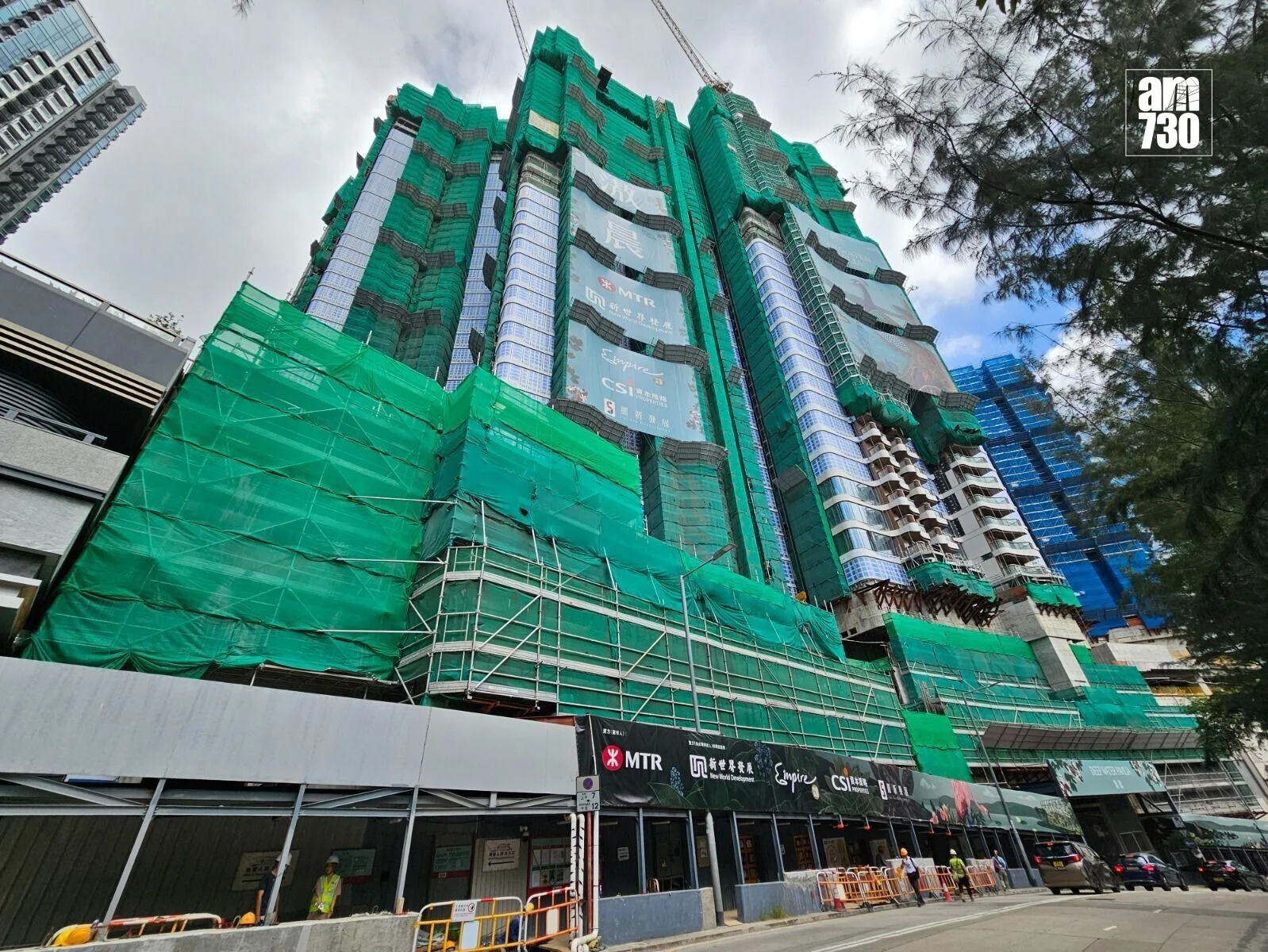 Tall high-rise under construction wrapped in green safety netting with scaffolding and cranes above, along a city street with barriers