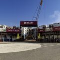 Entrance to a construction site with a tall crane and red Pavilia Rosa banners visible against a blue sky.