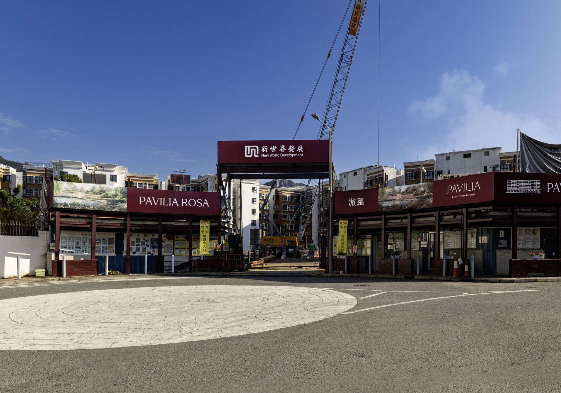 Entrance to a construction site with a tall crane and red Pavilia Rosa banners visible against a blue sky.