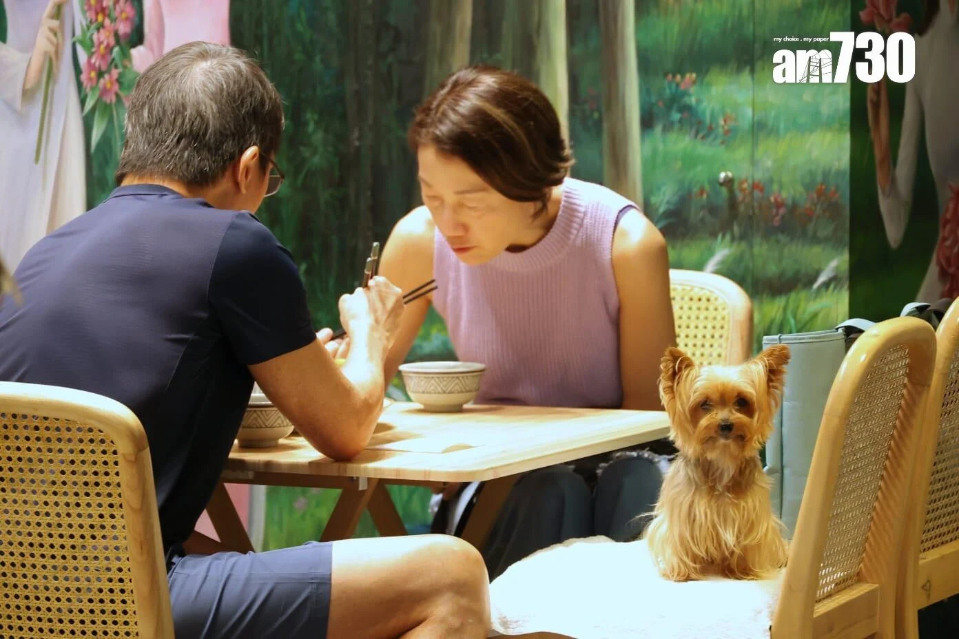 Couple dining at a wooden table while a small dog sits on a chair nearby, looking at the camera.
