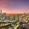 Panoramic city skyline at sunset with an orange to purple sky, dense high-rise buildings, and a foreground sports field.