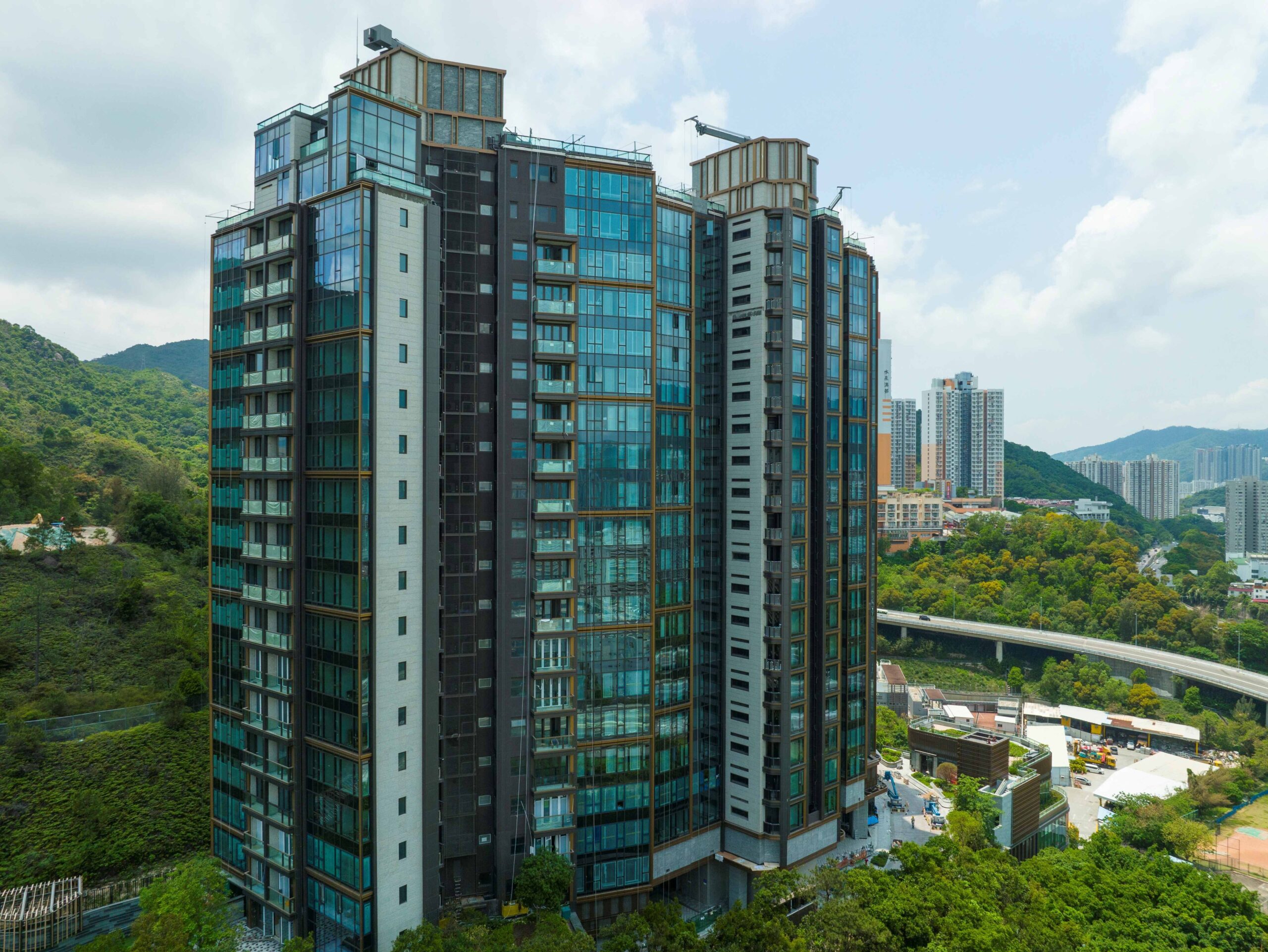 Modern glass-clad residential high-rise with balconies, set against a blue sky and surrounding hills and cityscape.