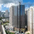 Skyline of high rise residential towers under a blue sky with scattered clouds a prominent glass clad central building dominates the scene with an elevated rail track at street level on the left and a city logo in the top right corner