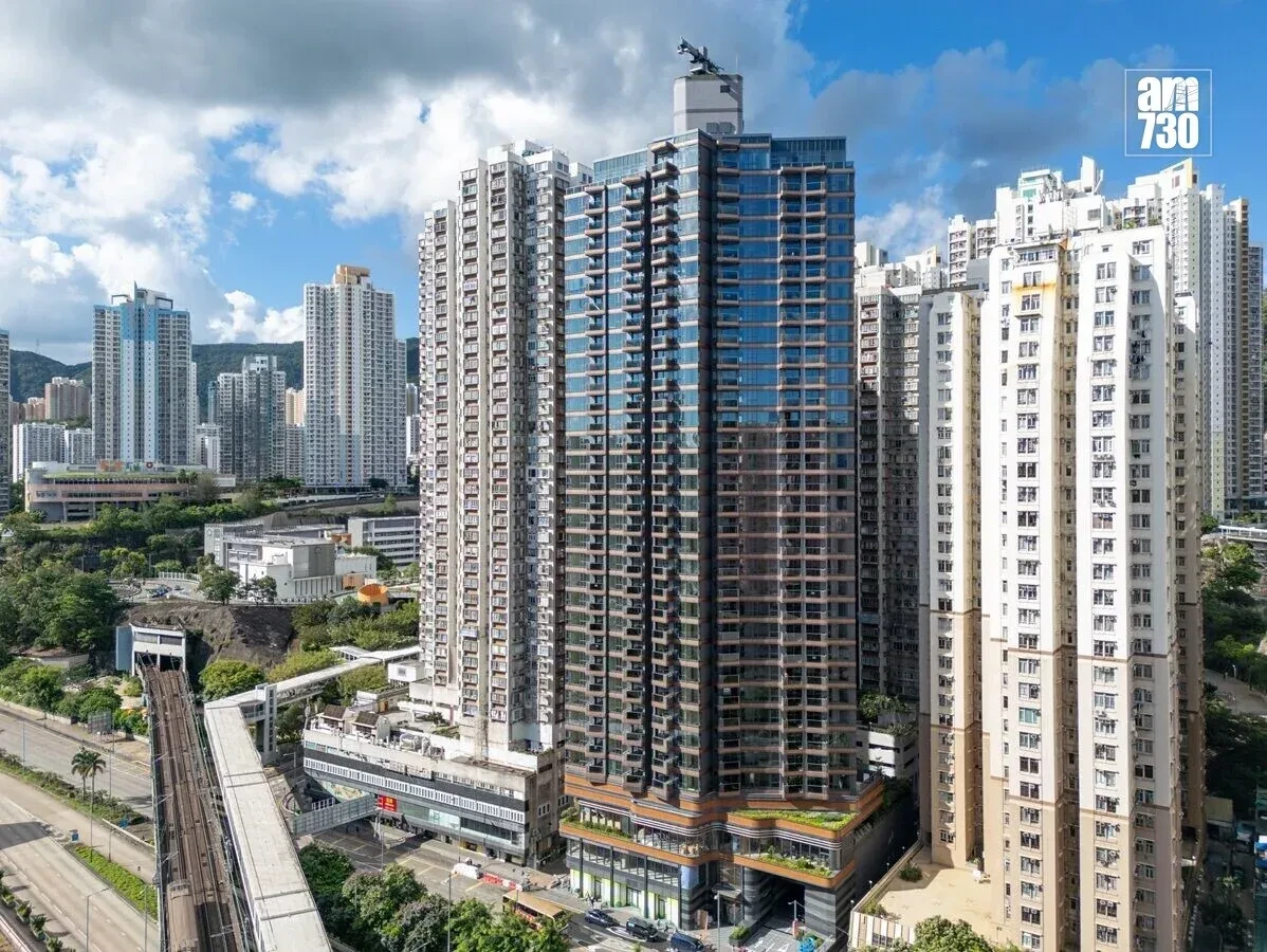Skyline of high-rise residential towers under a blue sky with scattered clouds; a prominent glass-clad central building dominates the scene, with an elevated rail track at street level on the left and a city logo in the top-right corner.